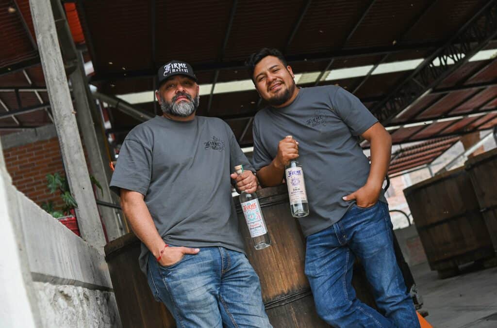 The brand owner and producer of Firme Mezcal stand in a palenque holding bottles